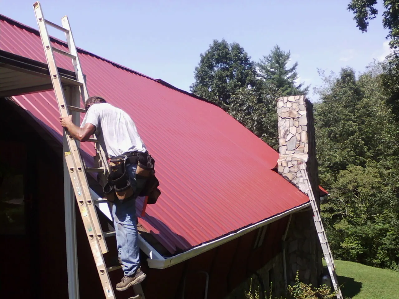 Roofer on a ladder beside a red metal roof and stone chimney — Soffit & Fascia Repair by Roofing Brothers in Barboursville